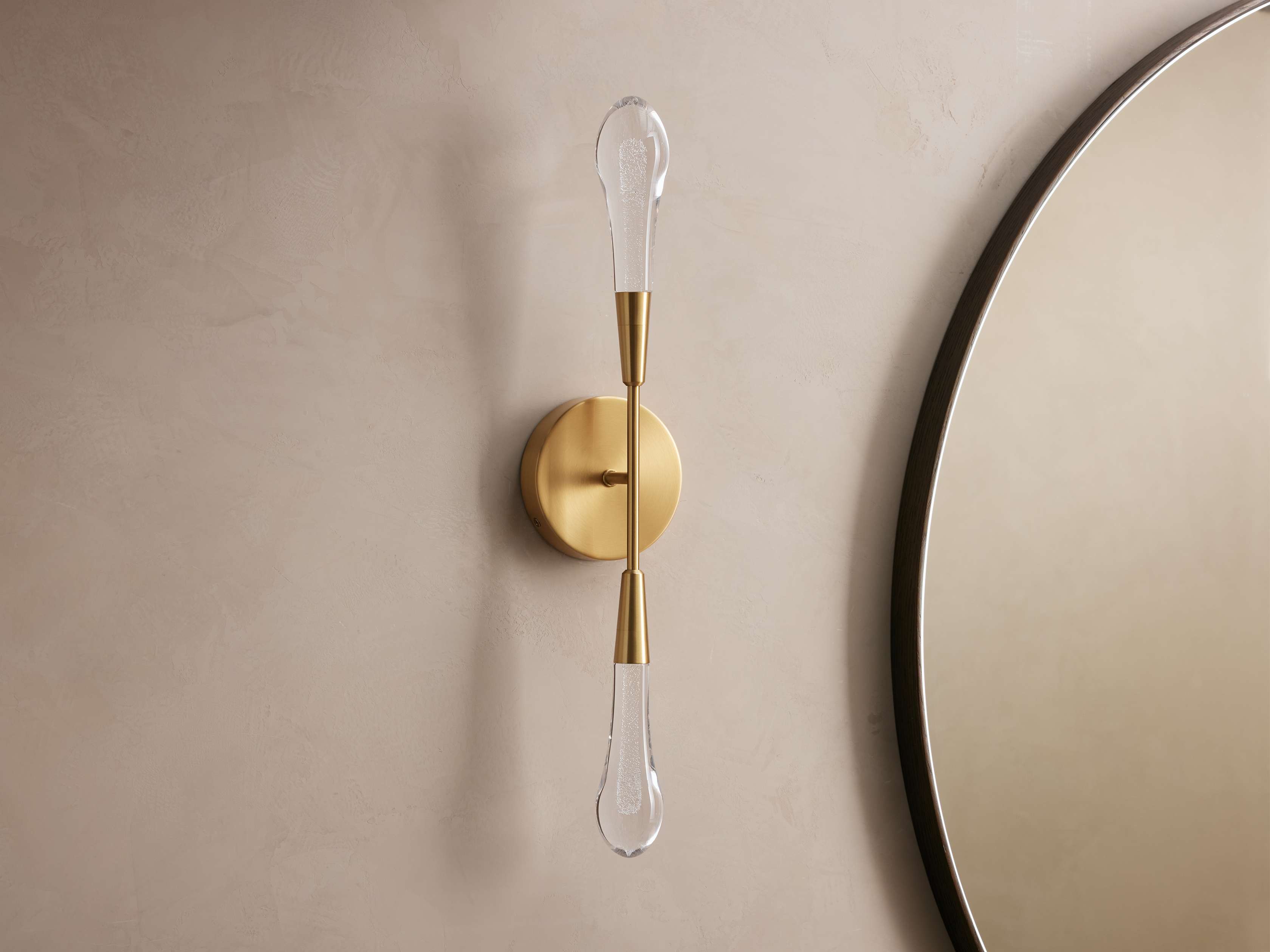 Gold wall sconce with clear glass shade on a beige wall next to a round mirror.